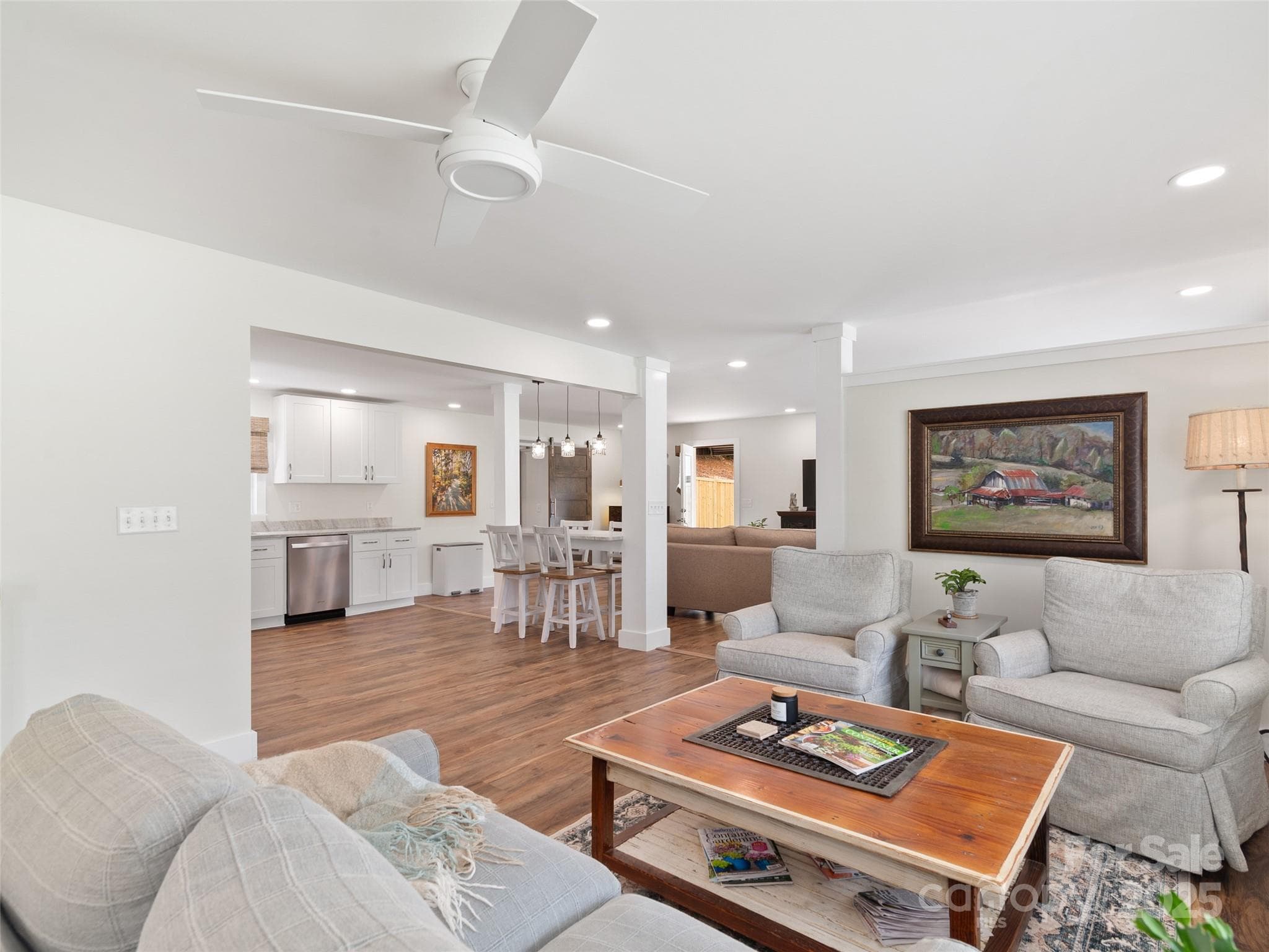 Bright open-concept living room with white ceiling fan, wooden coffee table, and adjacent kitchen area.