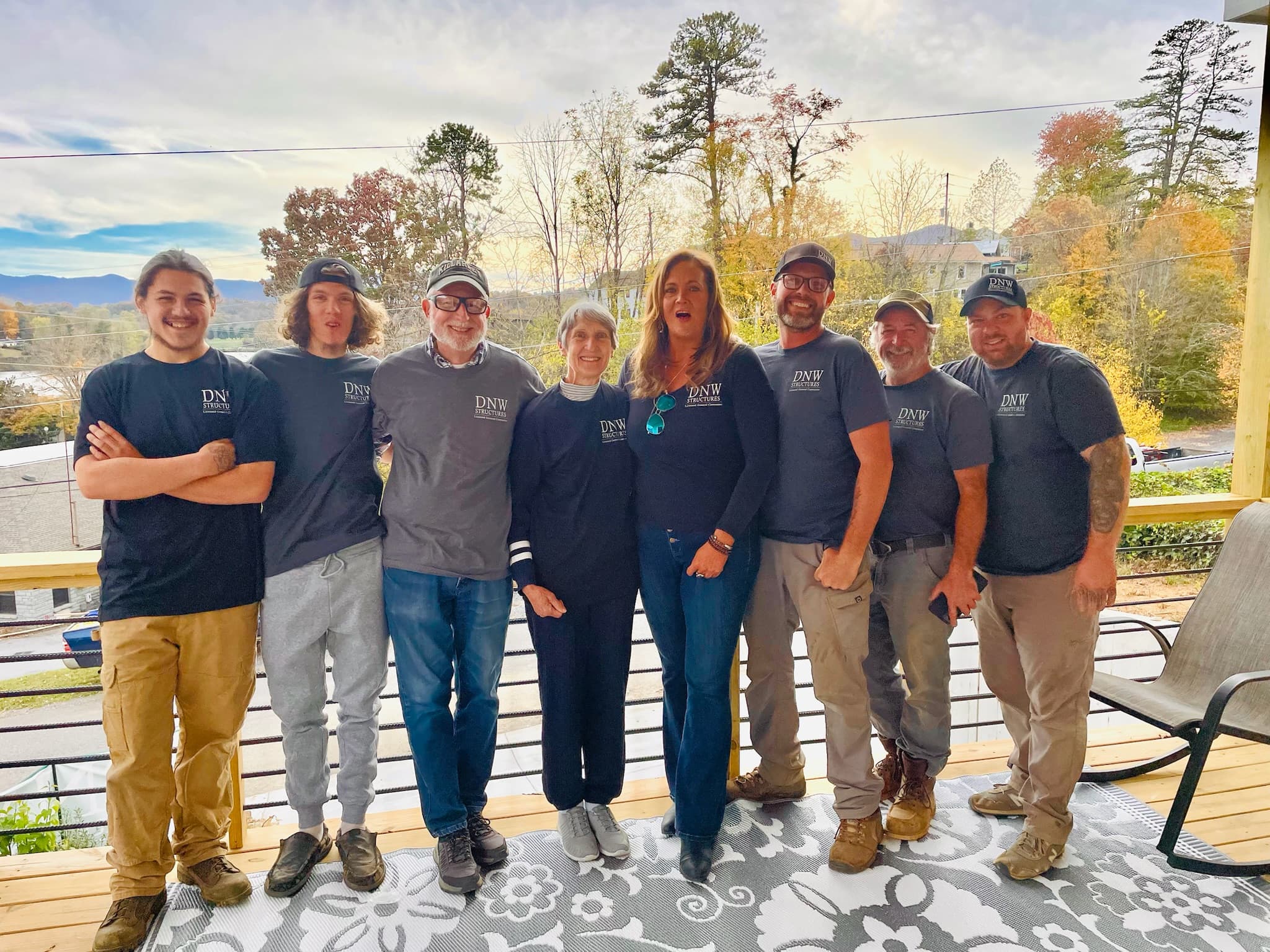 Eight people wearing DNW Structures shirts pose on a wooden deck with mountains behind them.