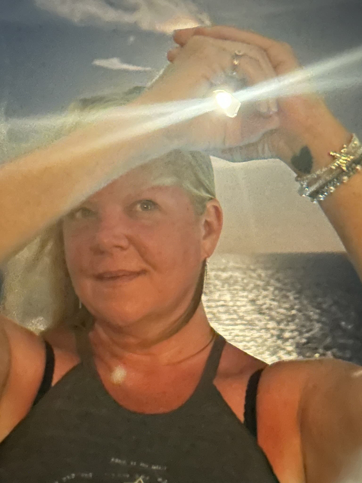 Smiling blonde woman holding hands overhead, framing a bright light flare against a shimmering ocean.